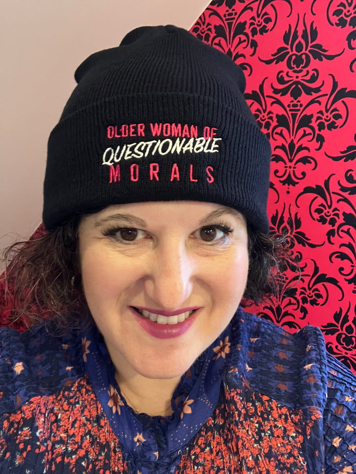 Deborah Wilde wearing black toque with pink and white text that reads “Older Woman of Questionable Morals”
